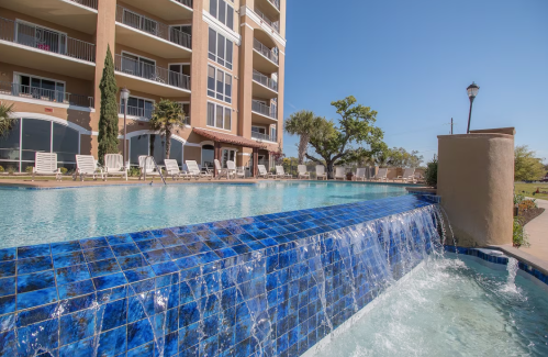 A serene pool with a blue tiled waterfall, surrounded by lounge chairs and palm trees, under a clear blue sky.