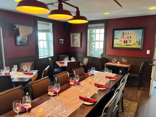 A cozy restaurant interior with a long table set for dining, featuring red walls, artwork, and warm lighting.