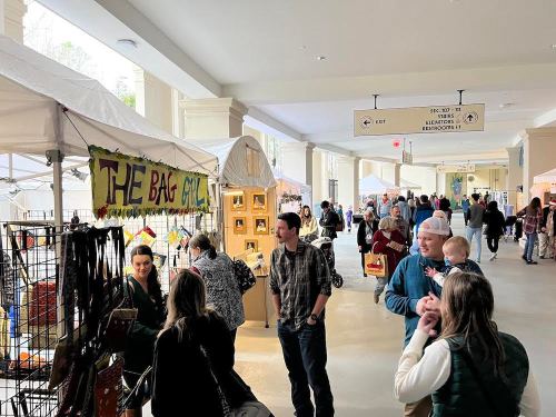 A bustling indoor market with vendors, shoppers, and displays of handmade goods under tents.
