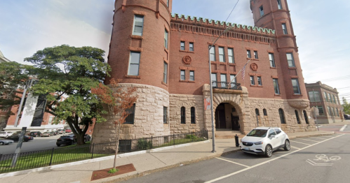 Historic brick building with a stone base, featuring towers and decorative elements, surrounded by trees and parked cars.