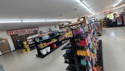 Interior of a store with shelves stocked with snacks and groceries, featuring a welcome sign and restrooms.