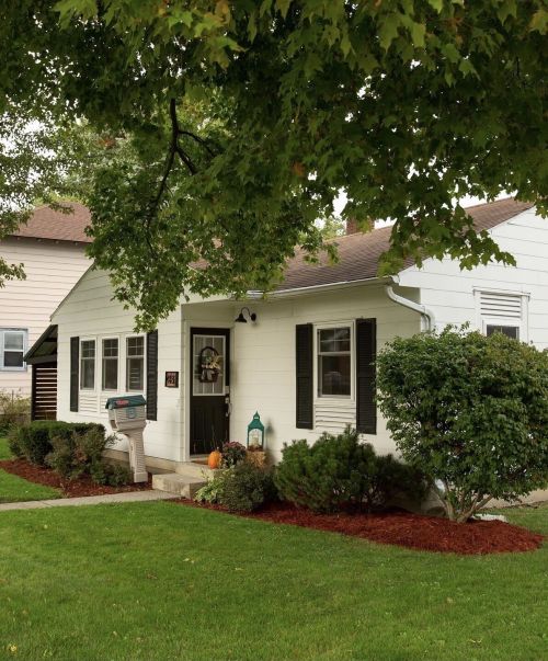 A charming white house with black shutters, a green lawn, and a mailbox in front, surrounded by shrubs and trees.