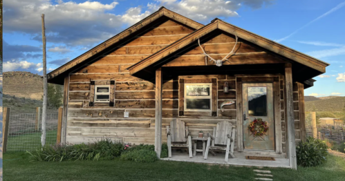A rustic wooden cabin with a porch, surrounded by grass and mountains under a blue sky with clouds.