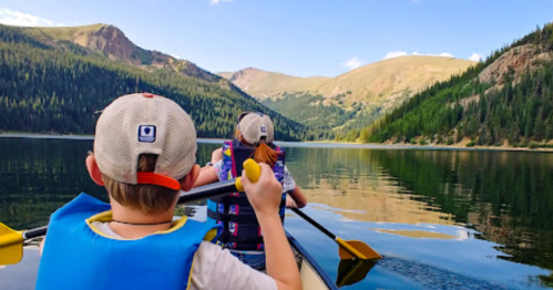 Two children paddle a canoe on a calm lake surrounded by mountains and trees under a clear blue sky.