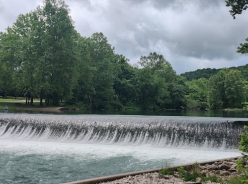 A serene landscape featuring a waterfall cascading into a calm river, surrounded by lush green trees under a cloudy sky.