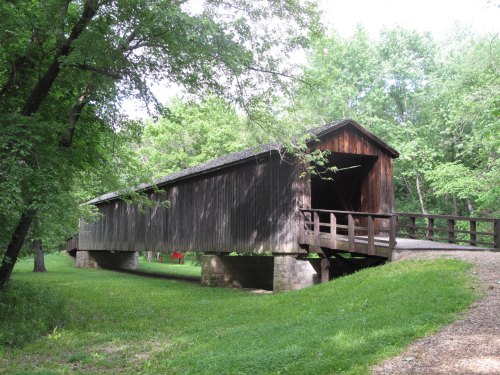 A wooden covered bridge surrounded by lush green trees and grass, with a gravel path leading to its entrance.