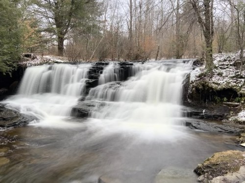 A serene waterfall cascades over rocks, surrounded by trees and a hint of snow in a tranquil forest setting.