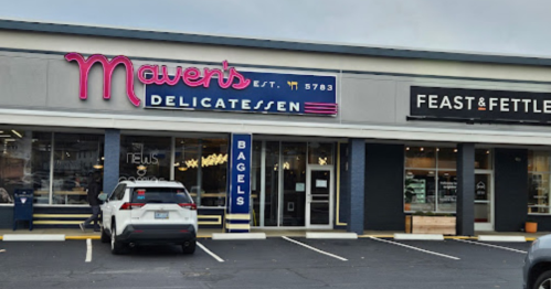 Exterior of Maven's Delicatessen with a sign and nearby Feast & Fettle, featuring parked cars in front.