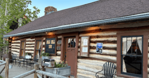A rustic log cabin inn with a sign, outdoor seating, and flower pots, set against a clear blue sky.