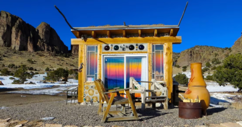 A colorful tiny house with a vibrant window, surrounded by snow and mountains, featuring outdoor seating and a fire pit.