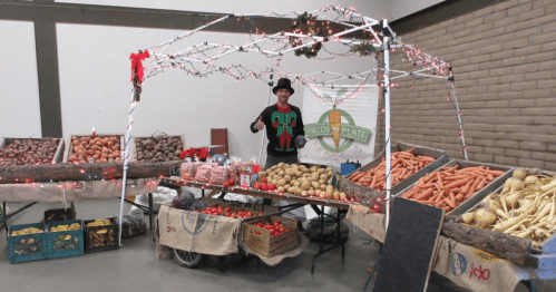 A festive market stall with a man in an elf costume, displaying various fresh vegetables and holiday decorations.