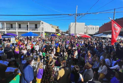 A large crowd gathers at a festive outdoor event, with colorful tents and decorations under a clear blue sky.