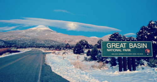 Snow-covered landscape with a road leading to a sign for Great Basin National Park under a blue sky.