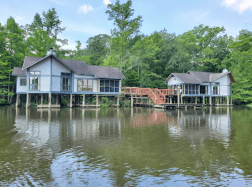 Two blue houses on stilts by a calm lake, surrounded by lush green trees and a wooden walkway connecting them.
