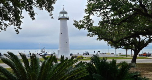 A white lighthouse stands by the shore, surrounded by palm trees and a cloudy sky, with cars parked nearby.
