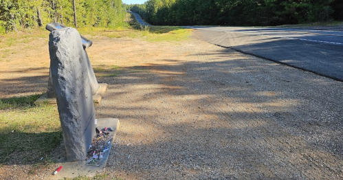 A weathered bench beside a gravel path, with a road visible in the background and trees lining the area.