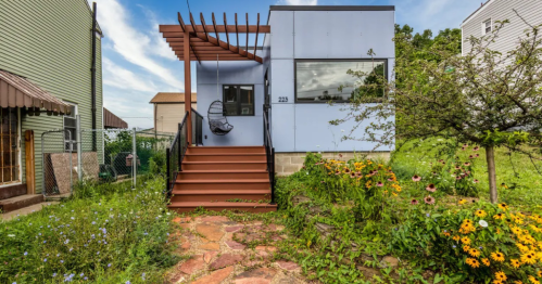 A modern house with a wooden porch, surrounded by greenery and colorful flowers under a blue sky.