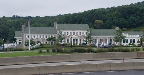 A large stone building with a green roof, surrounded by trees and grass, set against a hilly backdrop.