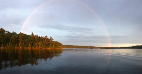 A serene lake scene with a rainbow arching over trees, reflecting on the calm water under a cloudy sky.