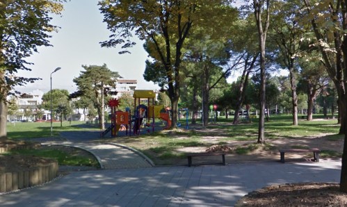 A colorful playground surrounded by trees in a park, with benches and grassy areas visible.