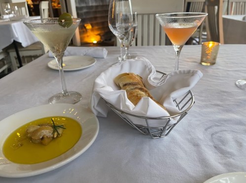 A table setting with two cocktails, a plate of olives in olive oil, and a basket of bread on a white tablecloth.