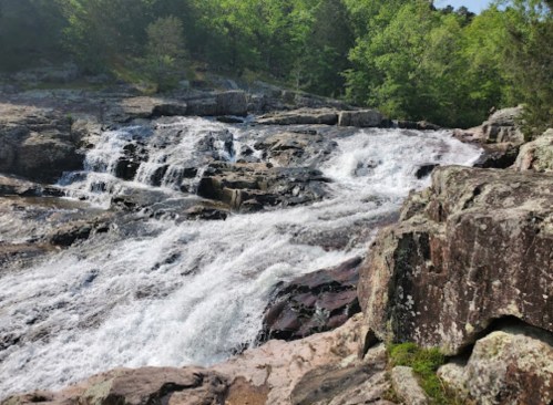 A cascading waterfall flows over rocky terrain, surrounded by lush green trees and a clear blue sky.