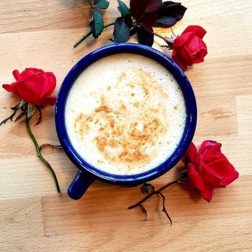 A blue mug of frothy coffee surrounded by red roses on a wooden surface.