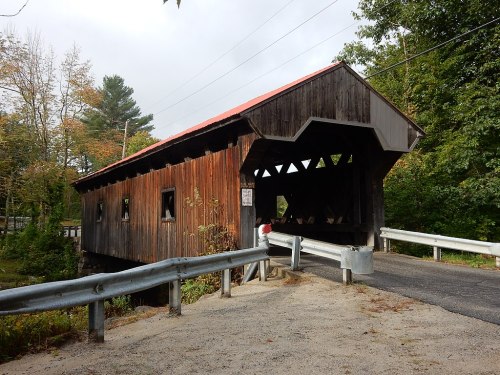 A rustic wooden covered bridge with a red roof, surrounded by trees and a gravel road.