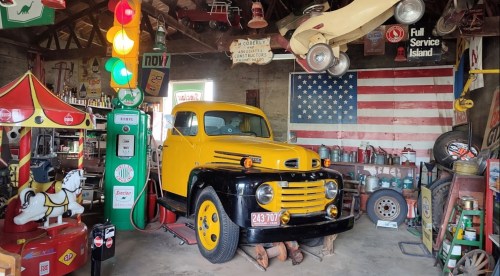 A vintage yellow and black truck in a rustic garage filled with retro signs, gas pumps, and Americana decor.