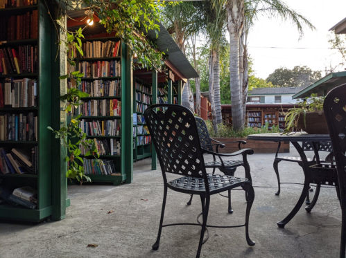 A cozy outdoor reading area with bookshelves, chairs, and palm trees in a tranquil setting.