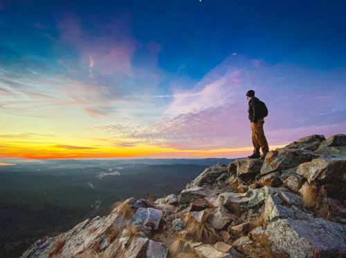 A person stands on a rocky outcrop, gazing at a colorful sunset over a mountainous landscape.