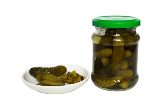 A jar of pickles with a green lid next to a white plate containing pickles and chopped pickled vegetables.