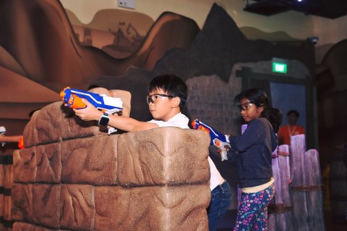 Two children play with toy blasters behind a foam barrier in a colorful indoor play area.