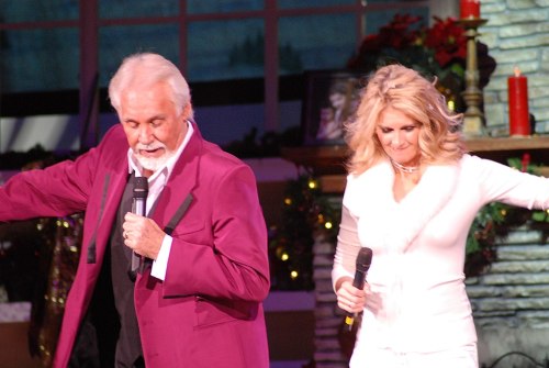 A man in a purple suit and a woman in a white outfit perform on stage, holding microphones, with festive decorations in the background.