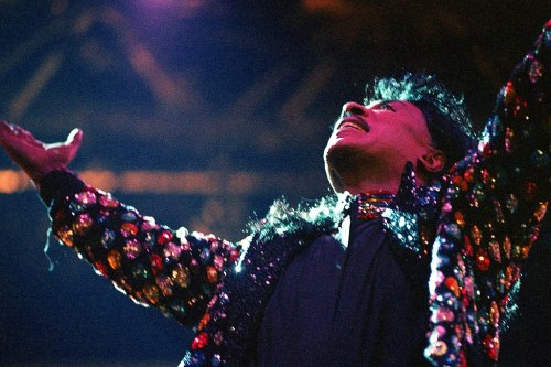 A performer in a sparkling, colorful outfit passionately gestures with arms raised, illuminated by stage lights.
