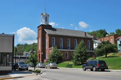 A brick church with a steeple stands beside a street lined with trees and parked cars under a blue sky.