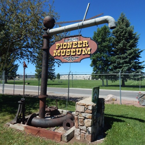 Sign for Pioneer Museum featuring a large pipe and vintage pump, set against a clear blue sky and green trees.