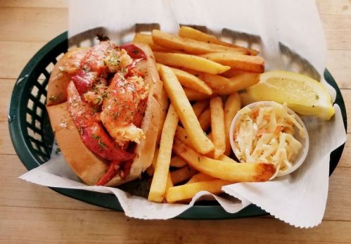 A lobster roll with fries and coleslaw, served with a lemon wedge in a green basket.