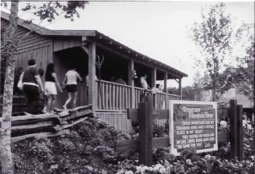 Black and white photo of a wooden building with people entering, surrounded by trees and flowers. Sign nearby reads about the location.