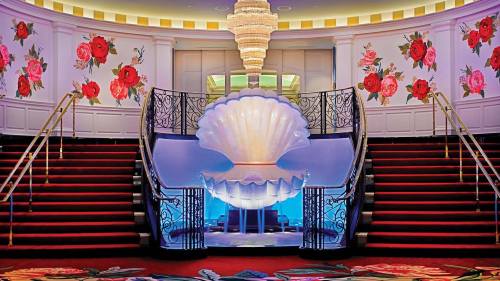 Elegant staircase with a large shell sculpture at the center, surrounded by floral wallpaper and a chandelier above.