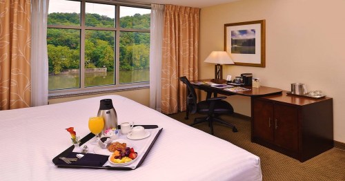 A cozy hotel room with a view of greenery, featuring a bed, desk, and a breakfast tray on the bed.