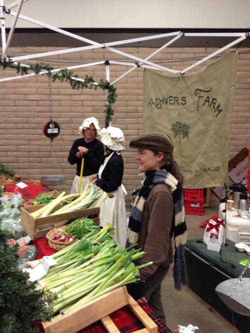 A farmer's market scene with fresh produce, two women in historical attire, and a man in a hat smiling at the stall.