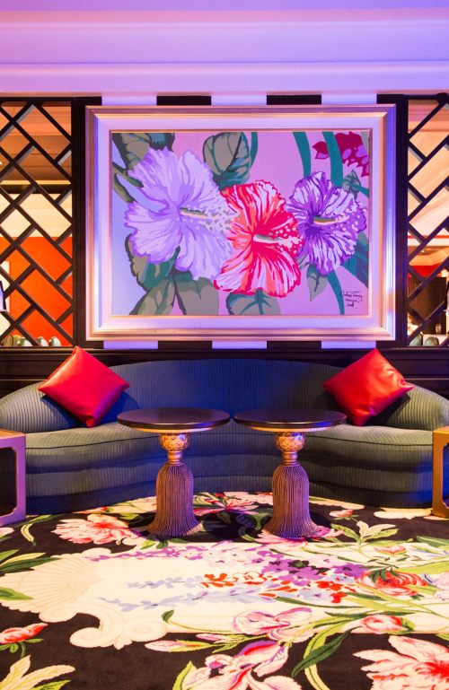 A stylish lounge area featuring a floral painting, a blue sofa with red cushions, and an ornate rug.