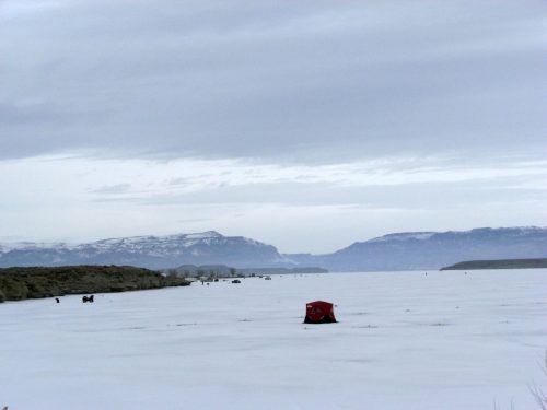 A red ice fishing shelter on a frozen lake, surrounded by snow-covered mountains under a cloudy sky.