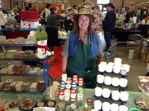 A smiling vendor in a straw hat stands behind a table displaying handmade soaps and candles at a craft fair.