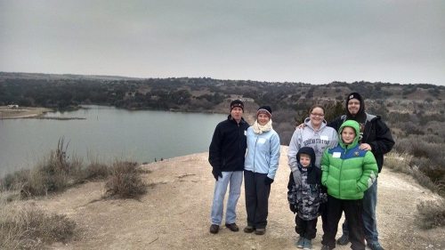 A group of six people stands on a hill overlooking a lake, with a cloudy sky and grassy landscape in the background.