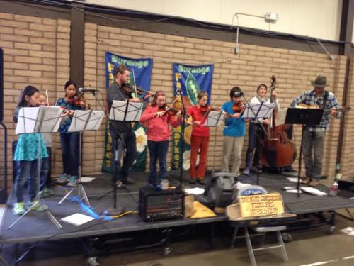 A group of musicians playing string instruments on stage, with sheet music and banners in the background.
