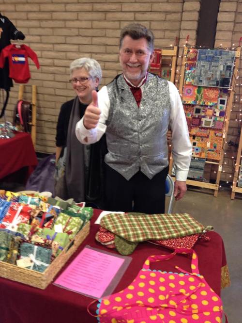 A man in a silver vest gives a thumbs up at a craft fair, with a woman smiling behind him and colorful items on the table.