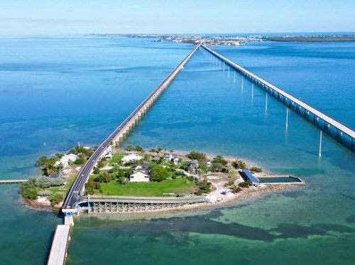 Aerial view of a long bridge extending over turquoise waters, leading to a small island with greenery and buildings.