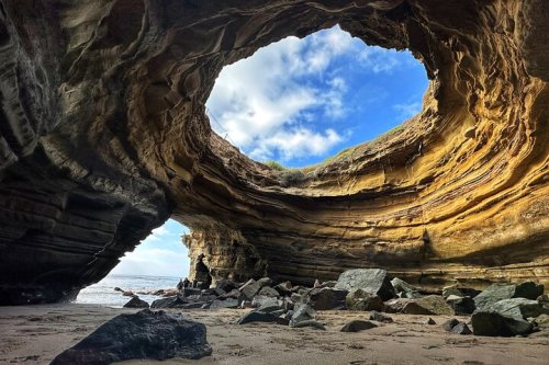 A rocky cave with a circular opening, revealing a blue sky and ocean beyond, surrounded by sandy beach and boulders.